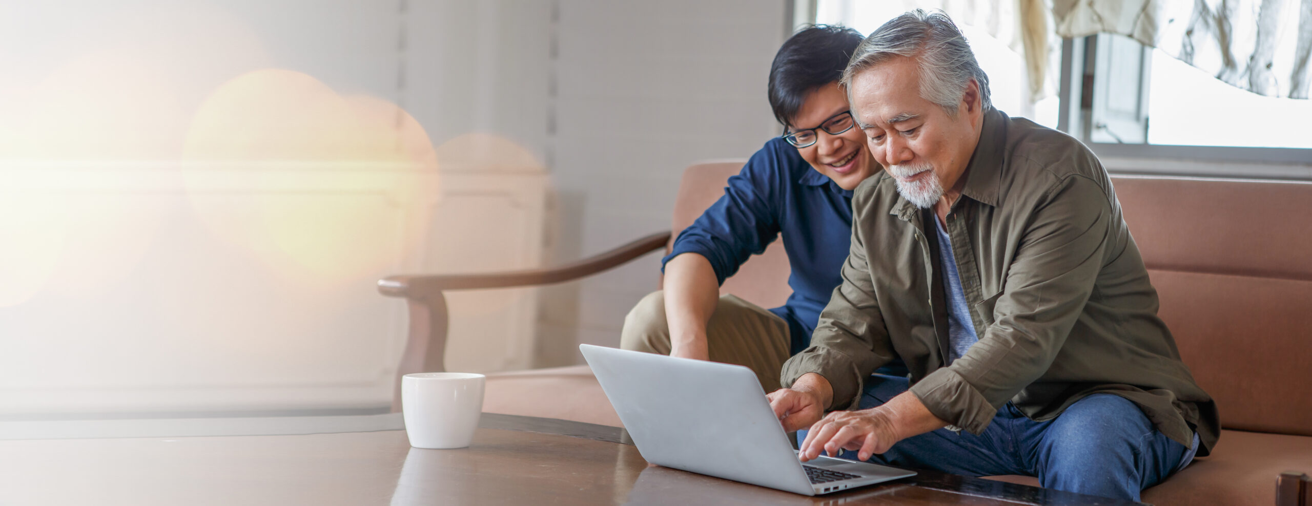 happy asian adult son and senior father sitting on sofa using laptop together at home considering Medicare enrollment periods