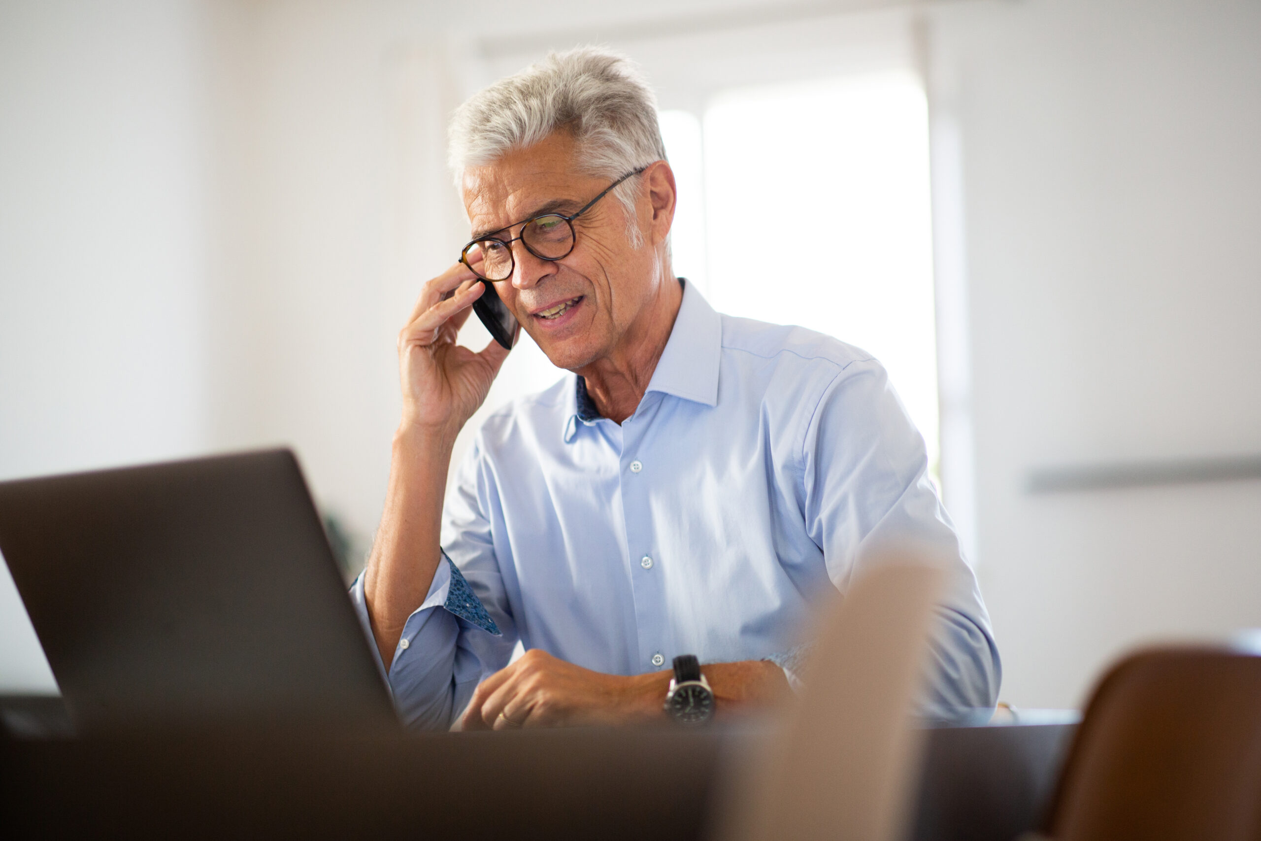 Older businessman sitting at office desk looking at laptop computer and talking on the phone with an agent comparing Medicare to group coverage